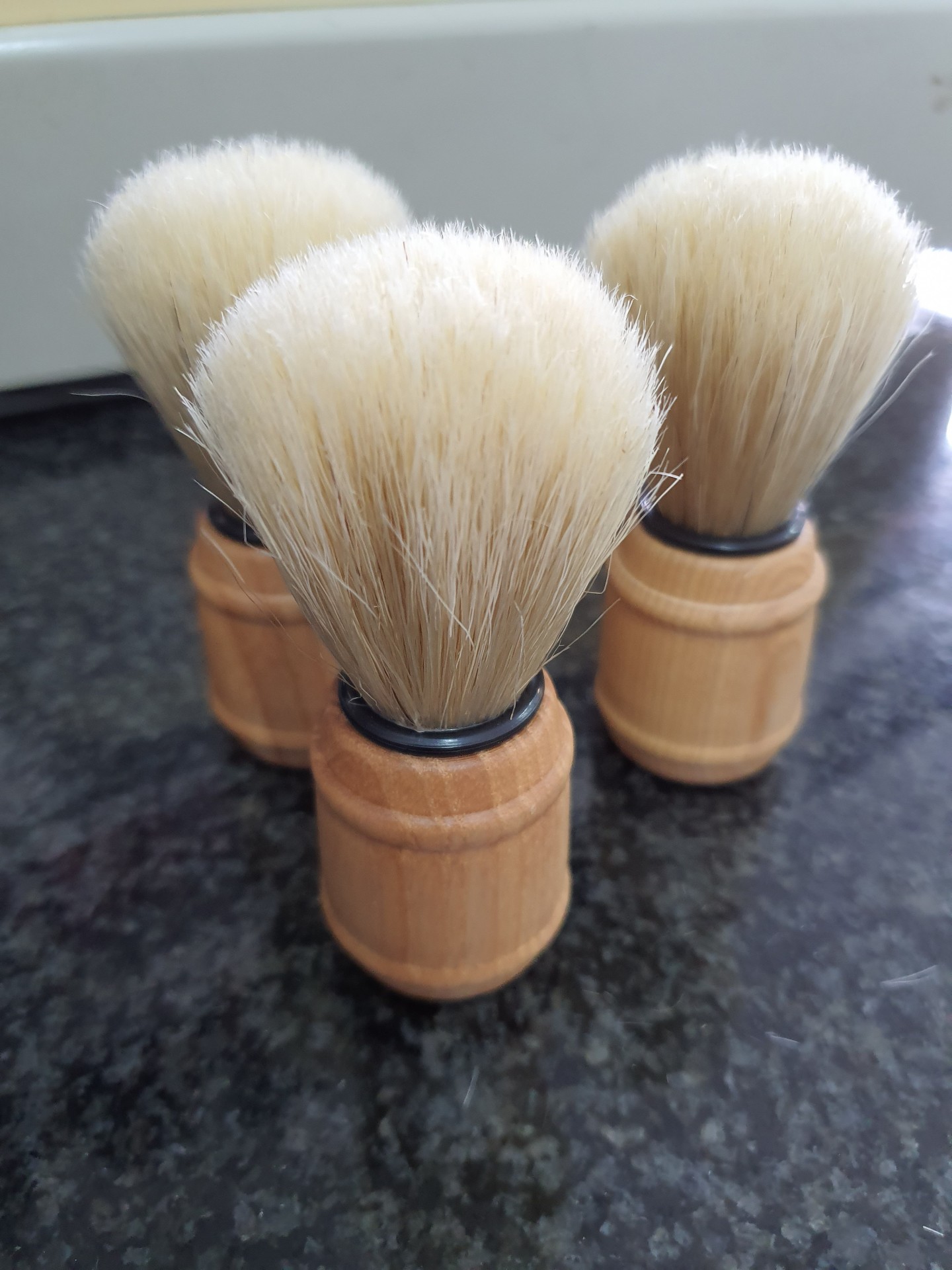 Três pincéis de barbear com cabos de madeira e cerdas brancas sobre superfície escura