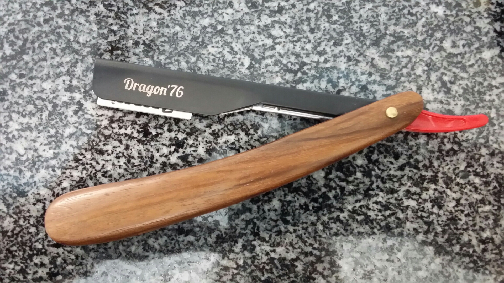 Barbeador naval preto com cabo de madeira castanha e proteção vermelha sobre granito cinza