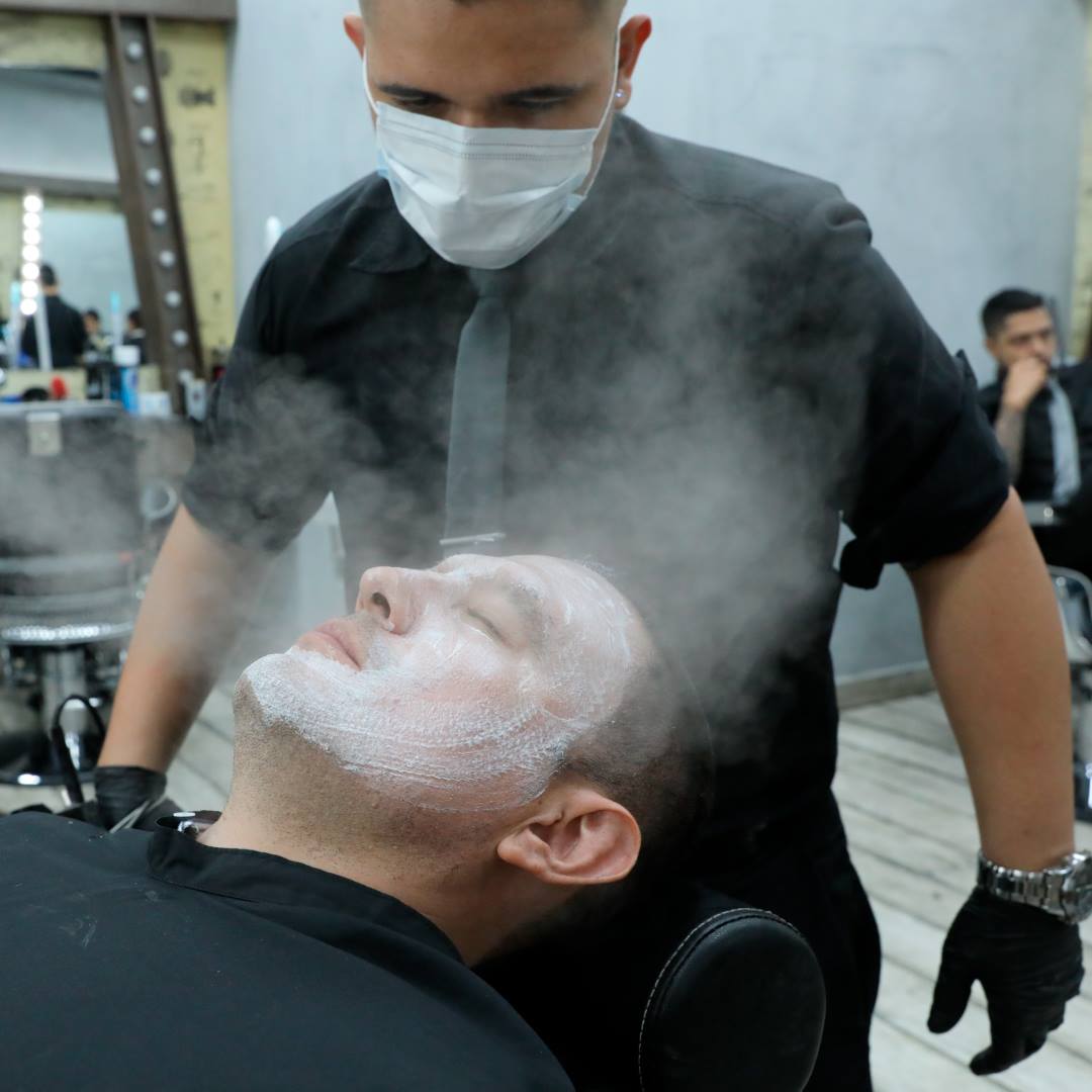 Homem a receber tratamento facial com espuma branca e vapor numa barbearia moderna com barbeiro de luvas pretas e máscara