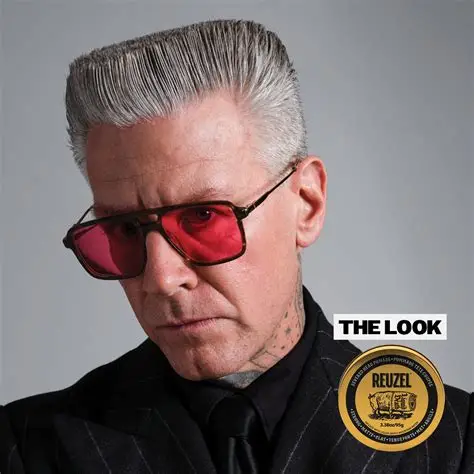 Homem com cabelo grisalho penteado, óculos de sol vermelhos, roupa preta e selo REUZEL