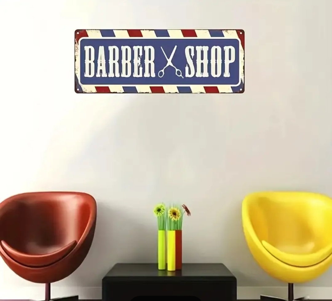 Placa decorativa 'BARBER SHOP' pendurada numa parede branca acima de duas cadeiras coloridas e uma mesa preta com vaso de flores.