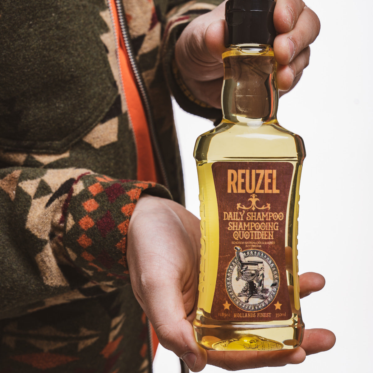 Frasco de shampoo REUZEL com rótulo castanho e amarelo segurado por uma pessoa