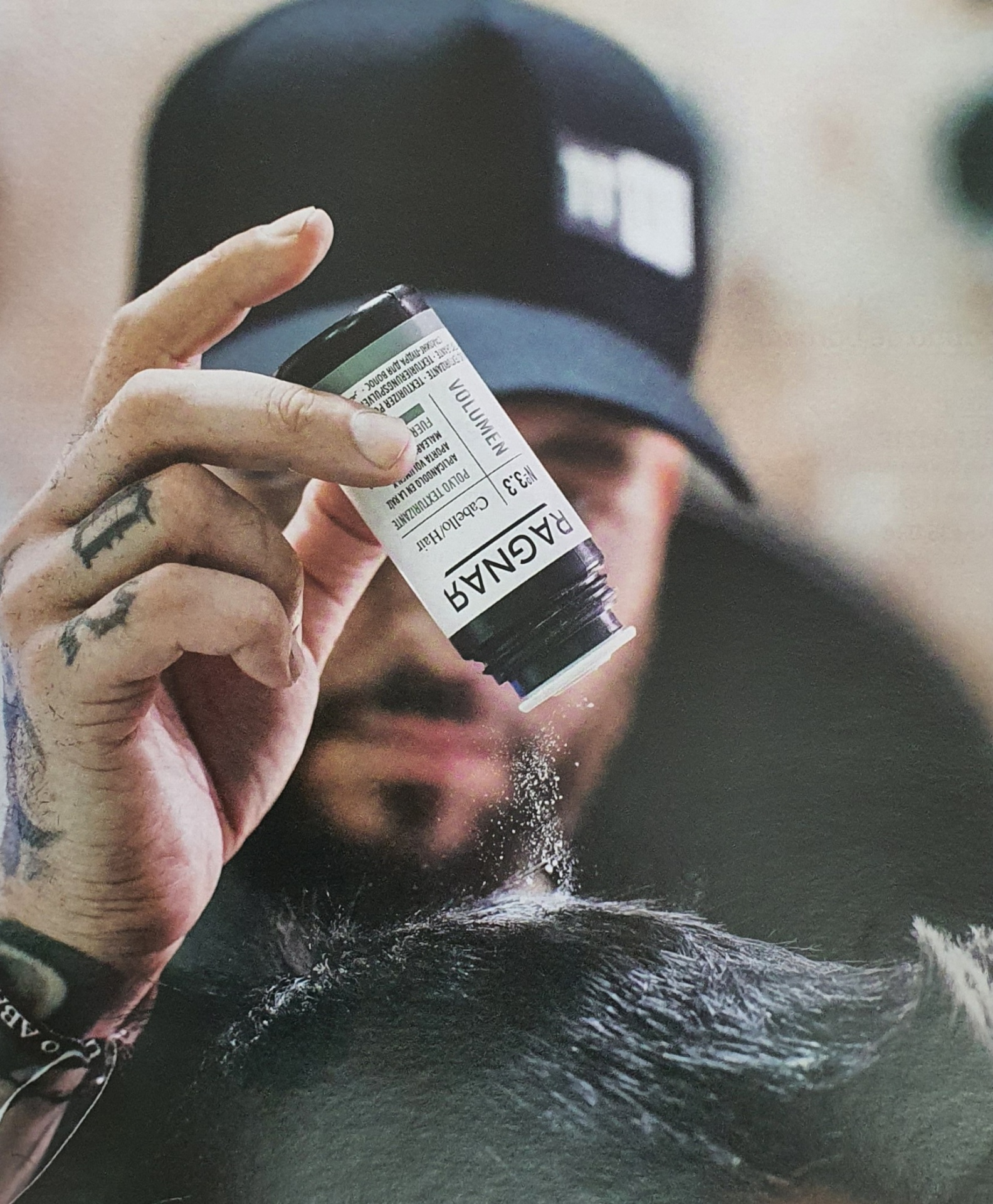 Frasco com pó a ser aplicado no cabelo de um cliente por uma pessoa com tatuagens e boné preto