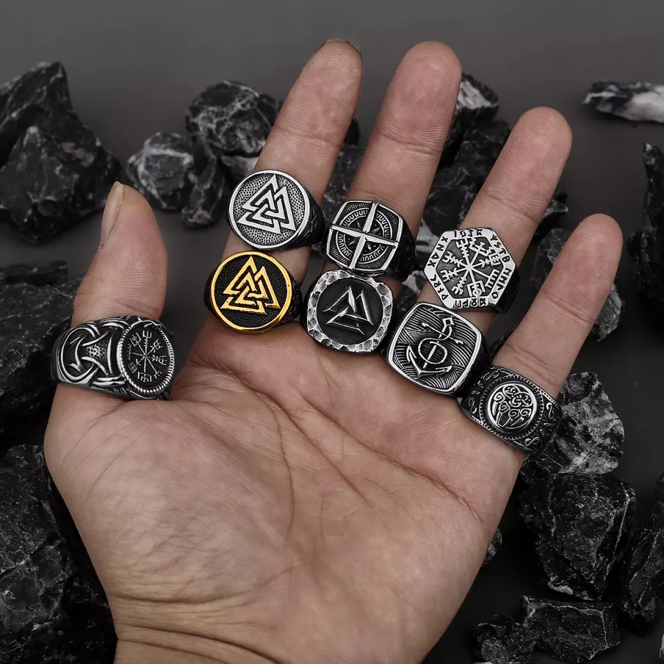 Mão com oito anéis de metal com símbolos nórdicos em fundo de pedras pretas e brancas.