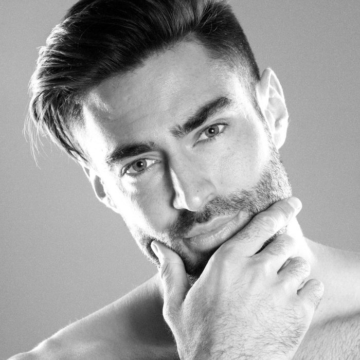 Homem com cabelo penteado e barba a tocar o queixo em fundo cinzento