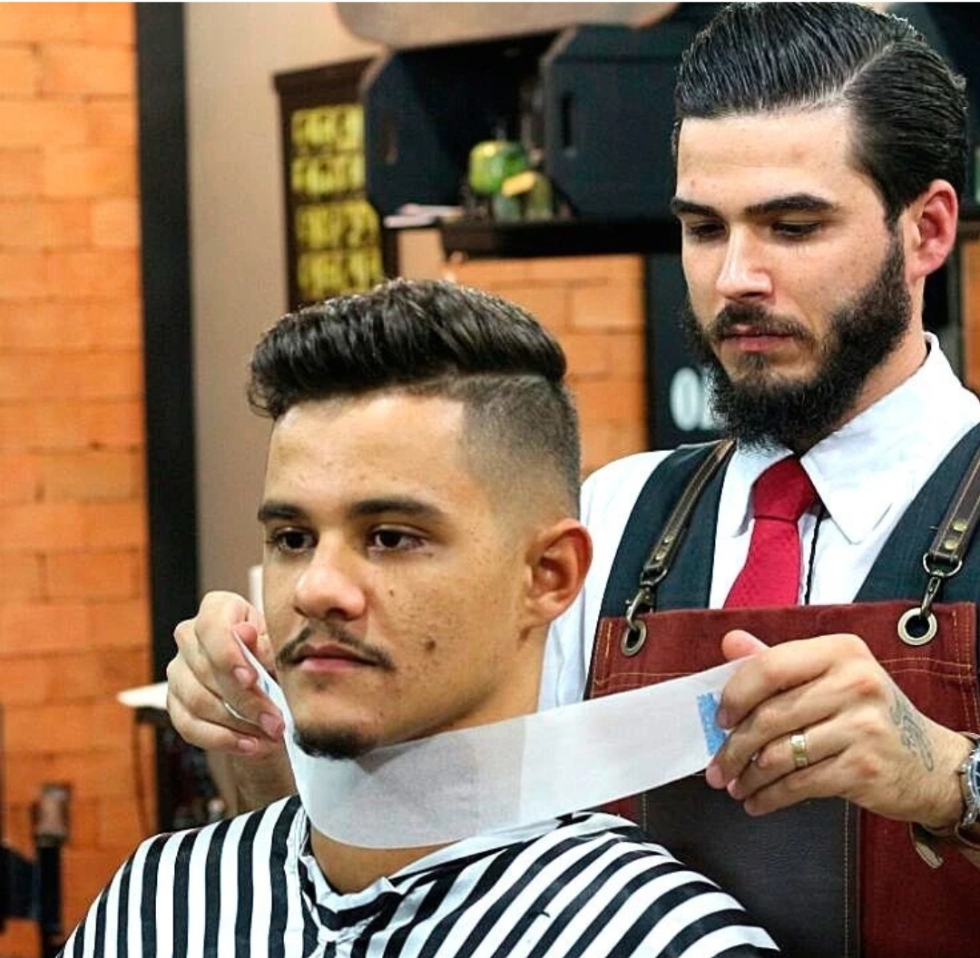 Barbeiro aplicando faixa de papel no pescoço de cliente com capa preta e branca num cabeleireiro