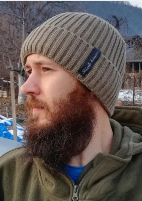 Homem de perfil usando gorro bege de malha com etiqueta preta e casaco verde em ambiente ao ar livre