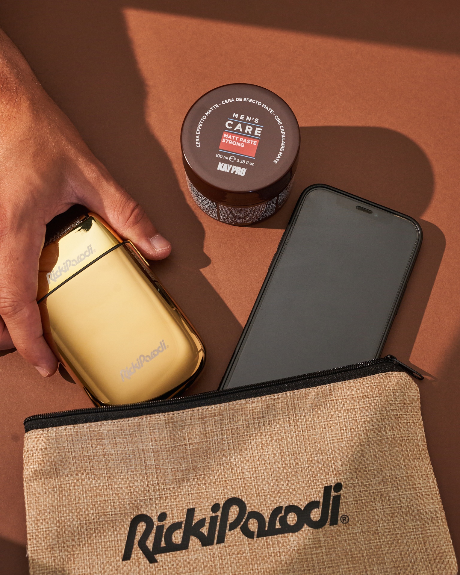 Frasco de pomada, estojo de tecido e dispositivo dourado com marca RickiParodi junto a um telemóvel sobre superfície castanha