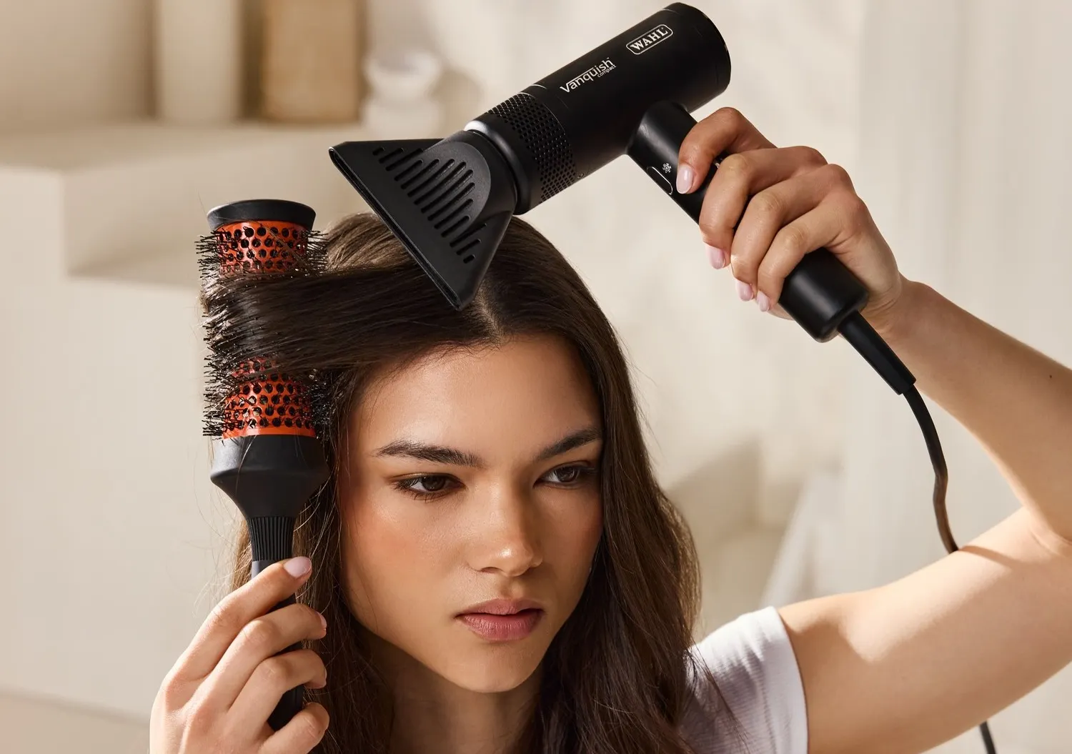Mulher a usar secador de cabelo Wahl Vanquish preto com escova redonda preta e laranja para alisar cabelo castanho.