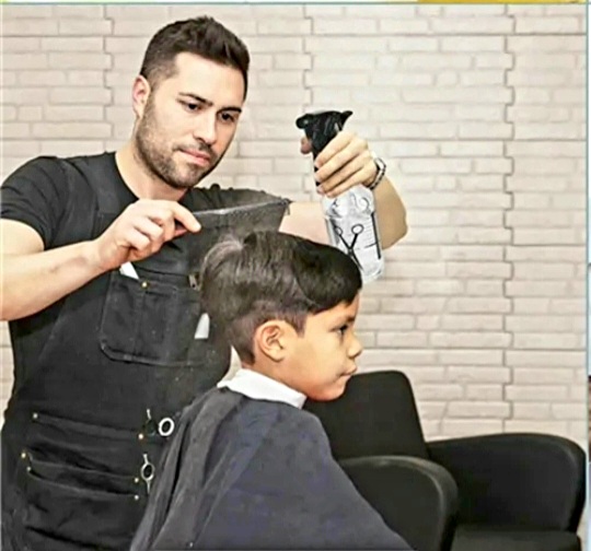 Barbeiro cortando cabelo de rapaz jovem com capa, usando tesoura e borrifador em ambiente com parede de tijolos brancos.