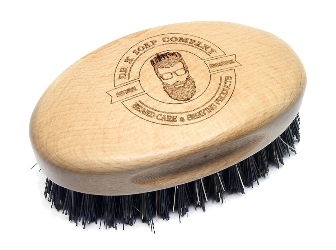 Escova para barba de madeira clara com cerdas pretas e logo gravado.