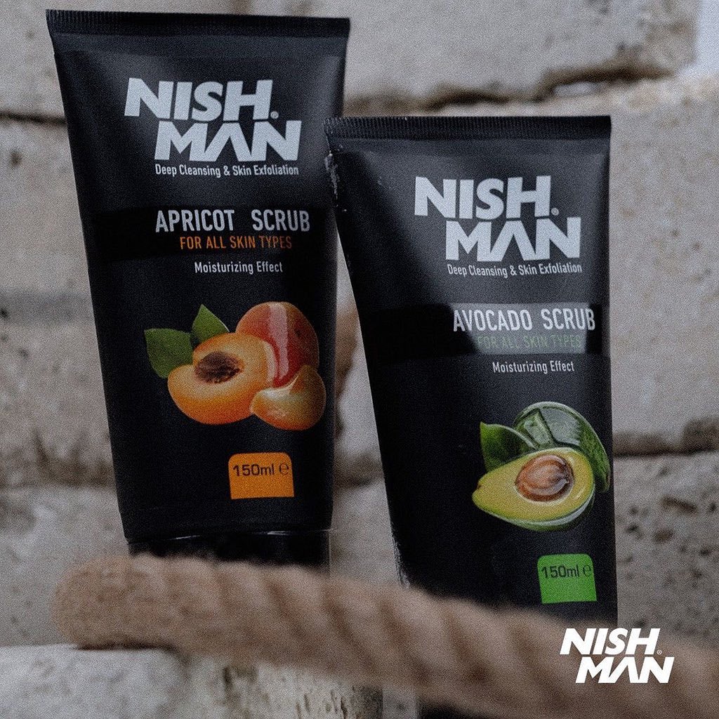 Dois tubos pretos de esfoliante facial NISH MAN Apricot Scrub e Avocado Scrub sobre superfície rústica com fundo de parede de pedra