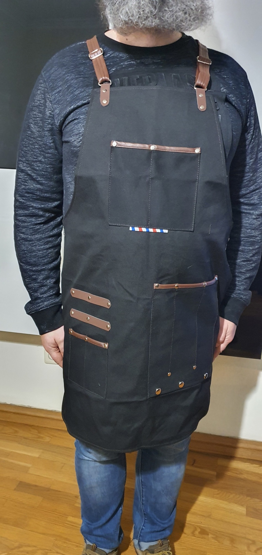 Avental preto com alças castanhas, vários bolsos e rebites metálicos usado por pessoa com camisola preta e jeans.