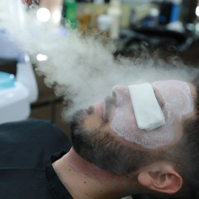 Homem com rosto coberto por creme e toalha nos olhos a receber vapor quente