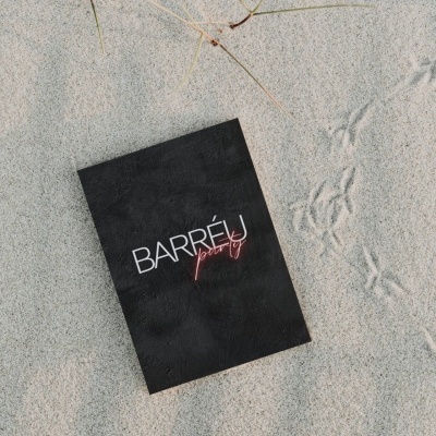 Caixa preta com texto 'BARRÉU party' na areia clara com relva seca