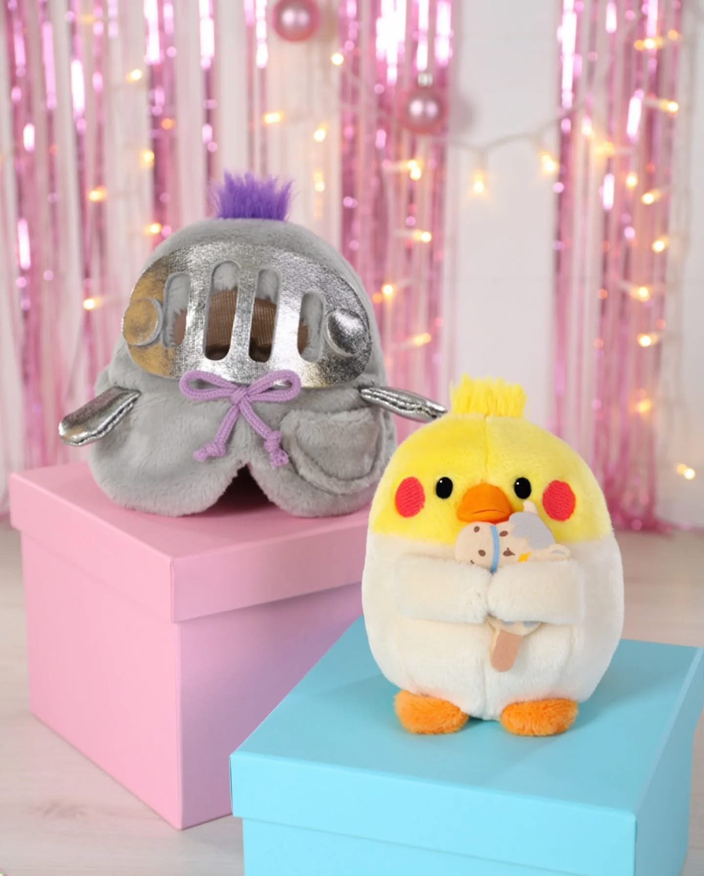 Peluches coloridos sobre caixas em cenário decorado com luzes e fitas cor-de-rosa.