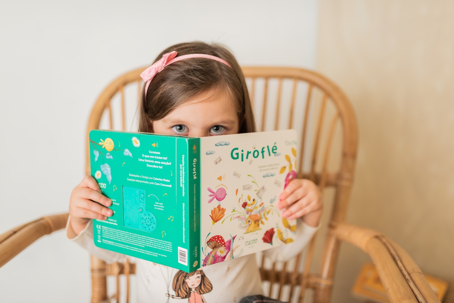 Criança a segurar um livro infantil 'Giroflé' com capa verde e branca, sentada numa cadeira de vime.
