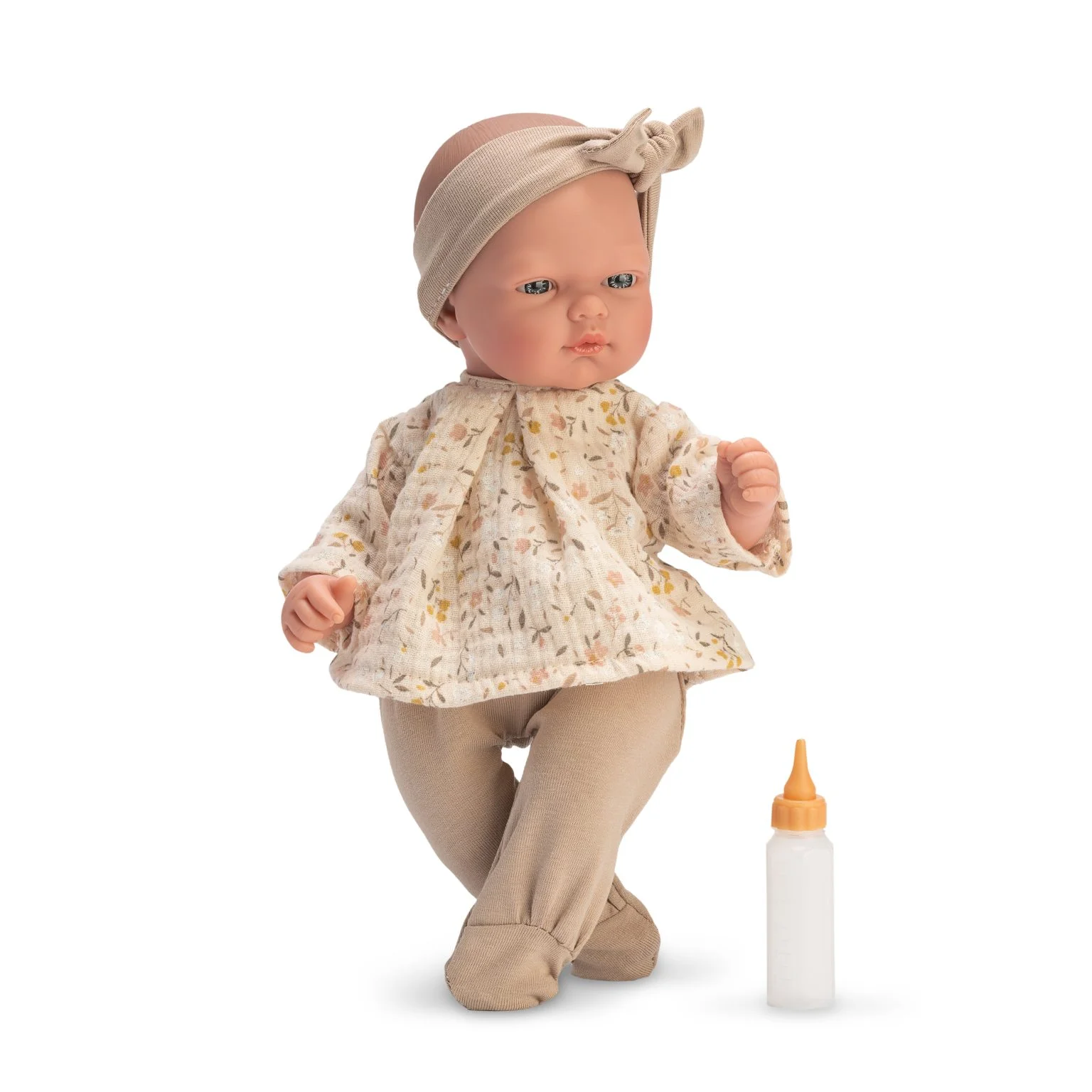 Boneca bebé com túnica floral e calças bege ao lado de um biberão