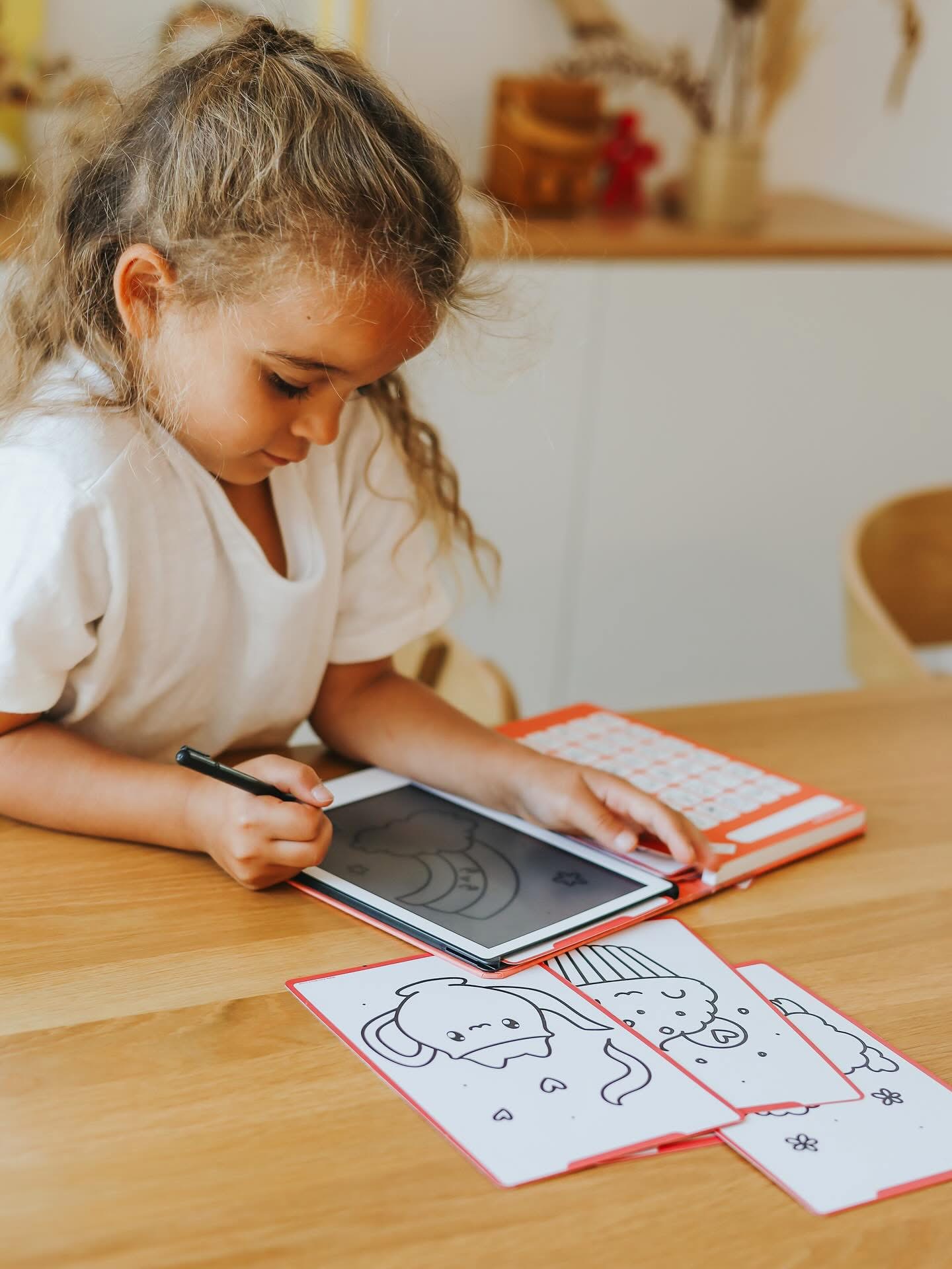 Criança a desenhar num tablet com capa laranja e teclado branco, mesa de madeira clara com desenhos infantis em preto e branco