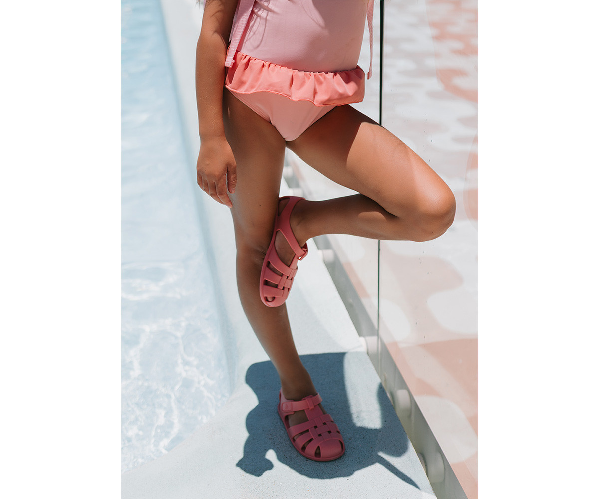 Criança com fato de banho rosa e sandálias rosa ao lado de piscina.