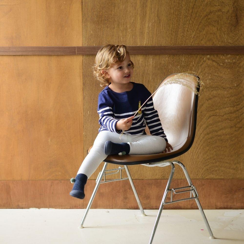 Criança sentada numa cadeira com estrutura metálica e assento claro junto a parede de madeira