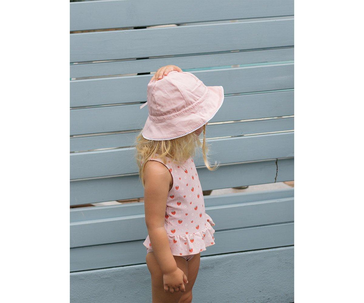 Criança de lado com swimsuit branco de cerejas e chapéu rosa claro, frente a parede azul