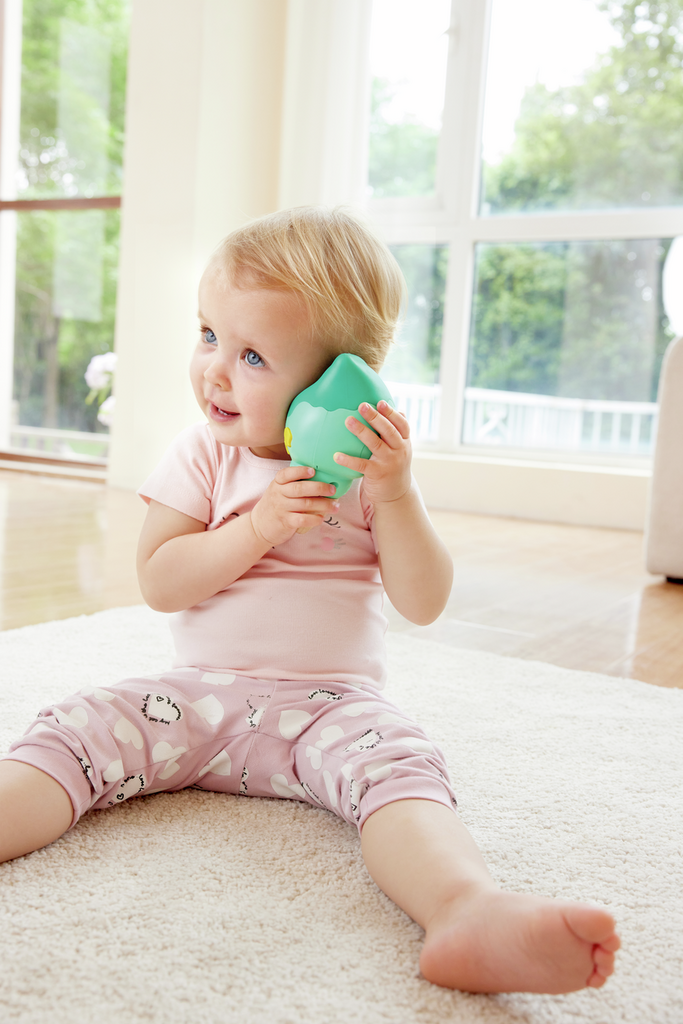 bebé vestido de pijama rosa sentado com brinquedo verde em tapete claro