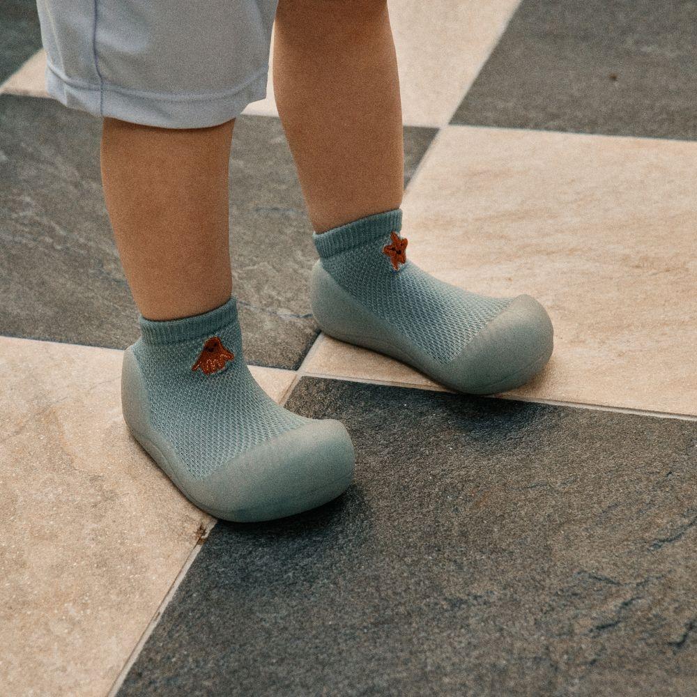 Calçado infantil azul claro com textura de malha e bordado castanho em piso xadrez