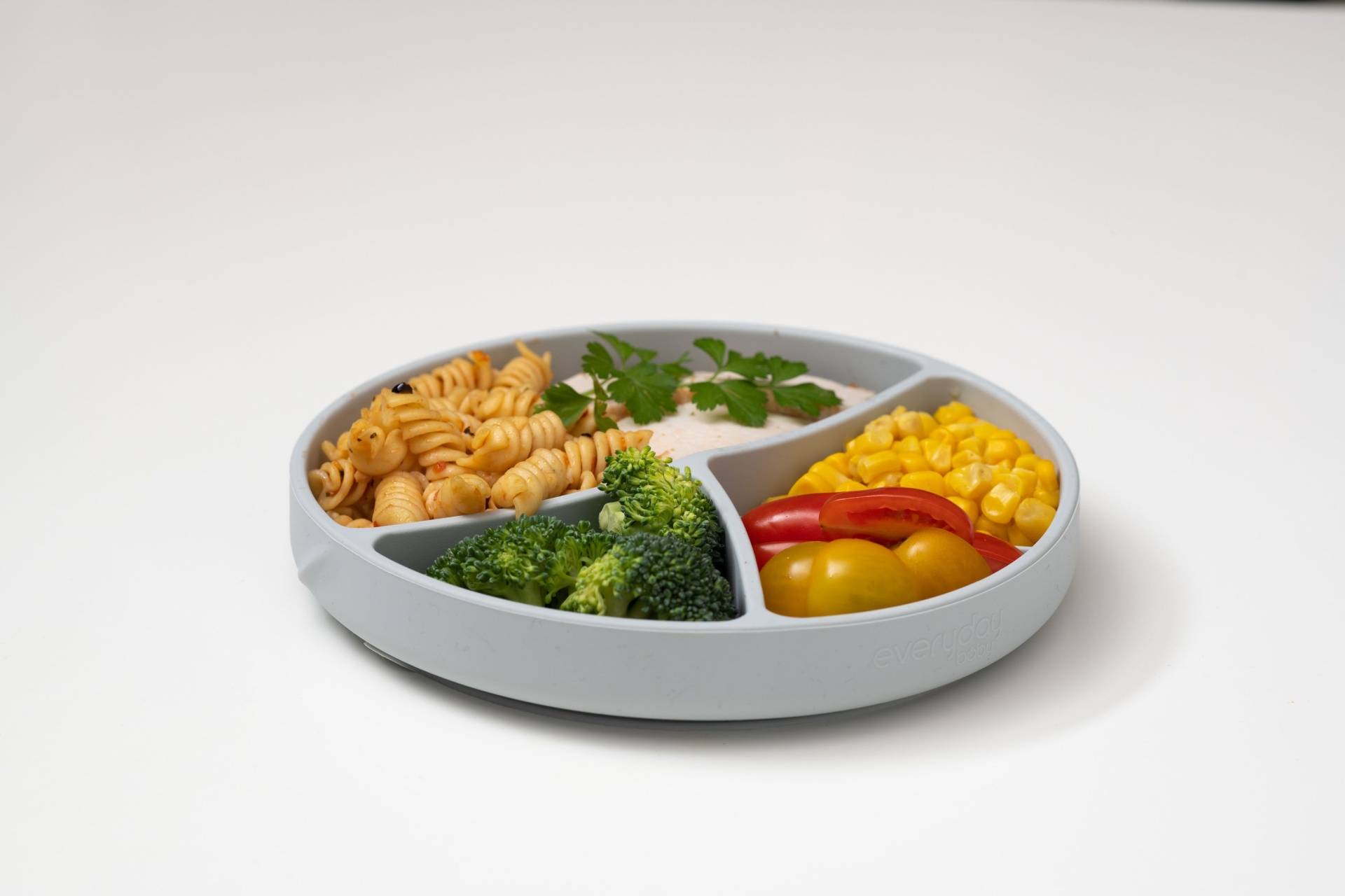 Prato redondo compartimentado com fusilli, vegetais variados numa superfície branca