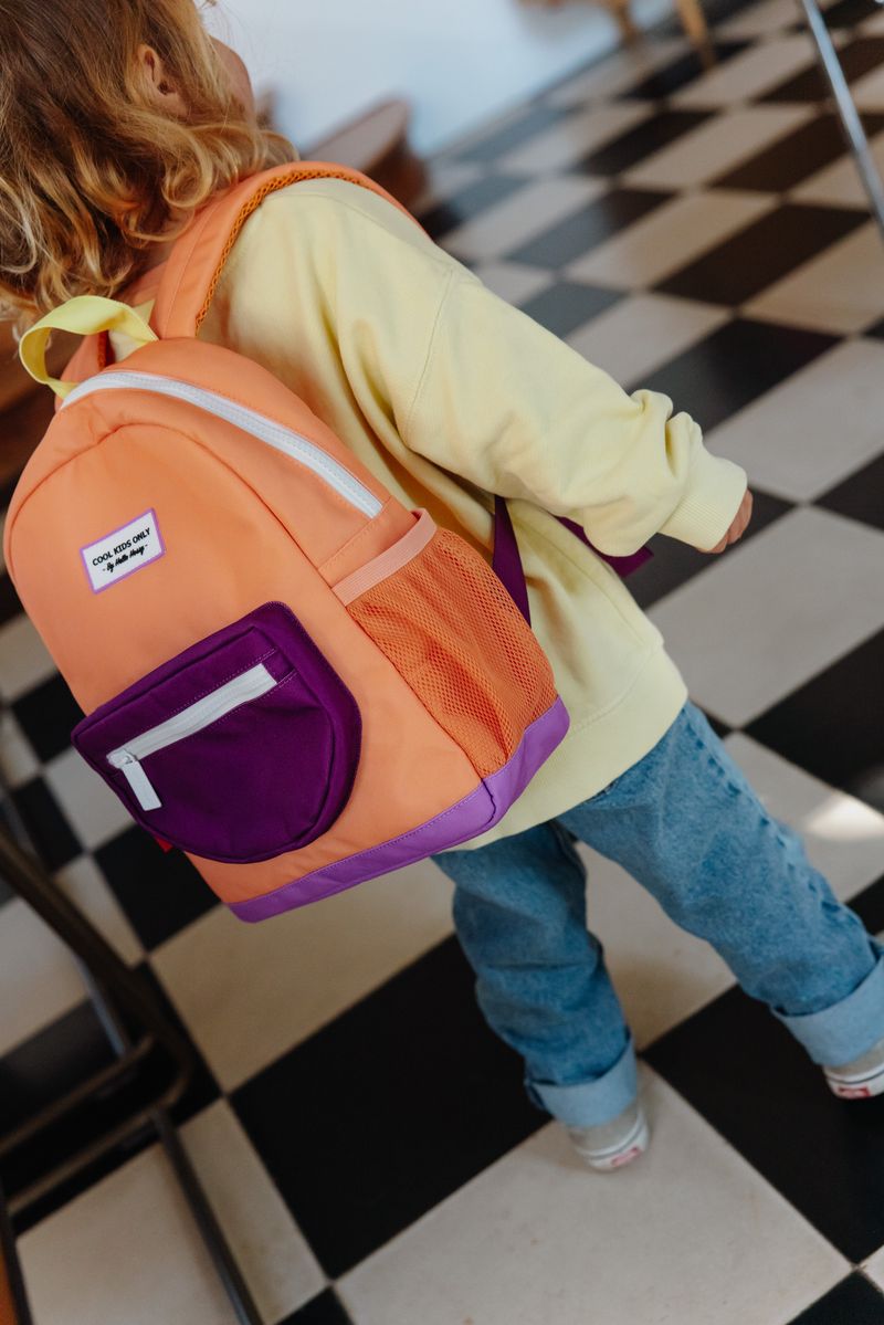 Mochila escolar laranja e roxa com bolso frontal roxo, usada por criança com casaco amarelo e calças azuis em piso xadrez.