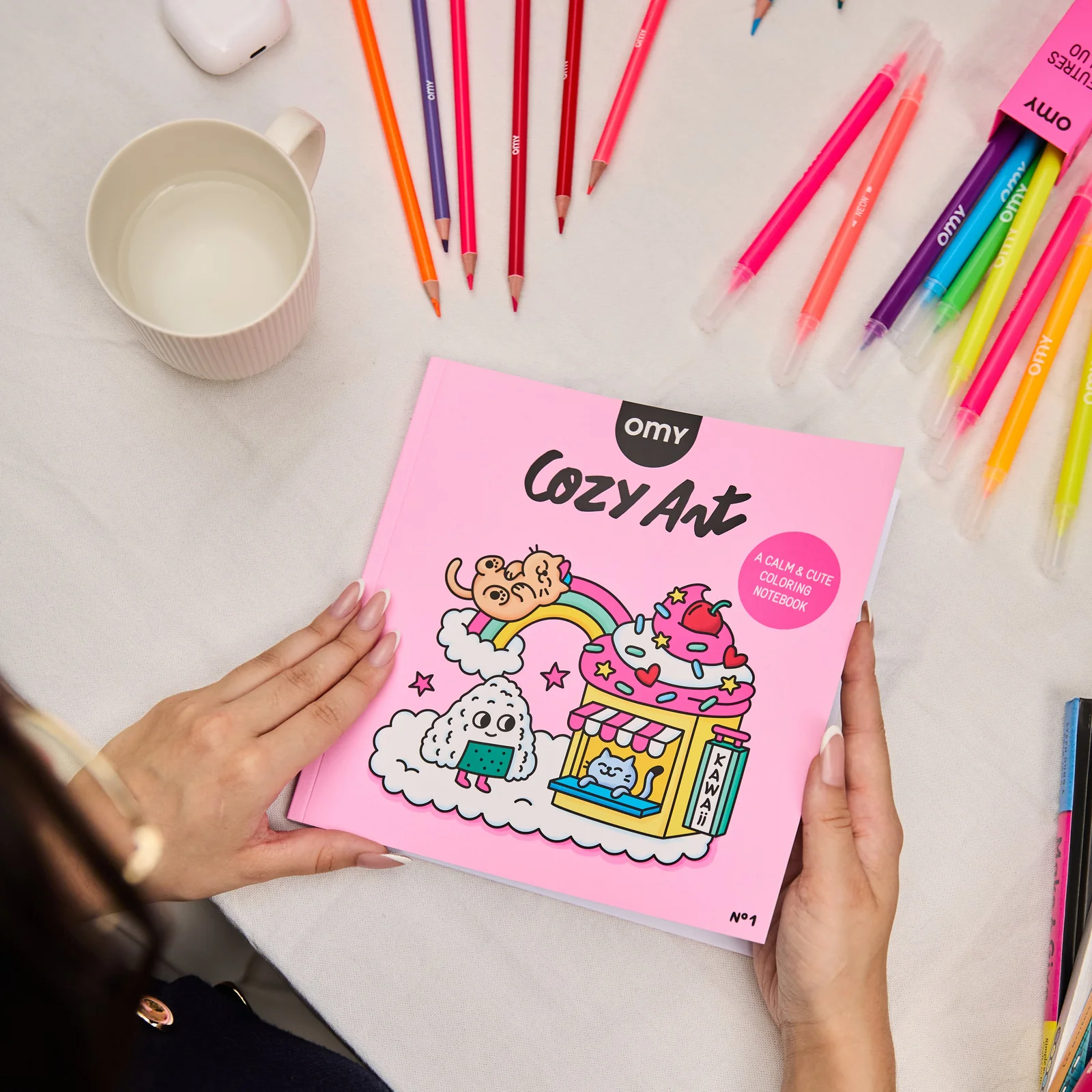 Livro de colorir Cozy Art rosa com desenhos kawaii e lápis e marcadores coloridos à volta.