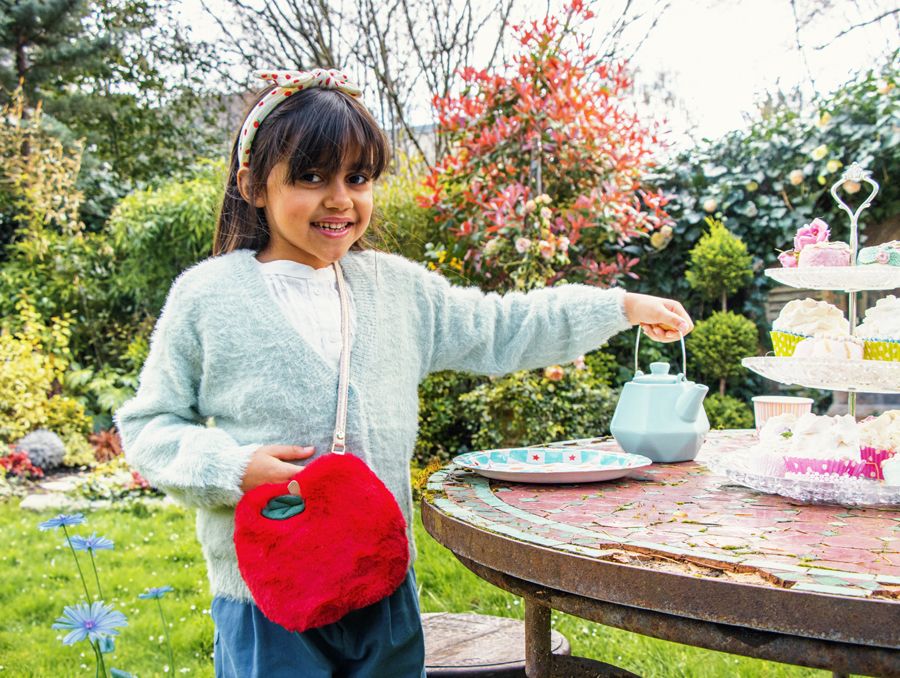 Menina no jardim com regador azul e bolsa vermelha peluda junto de mesa com bolo.