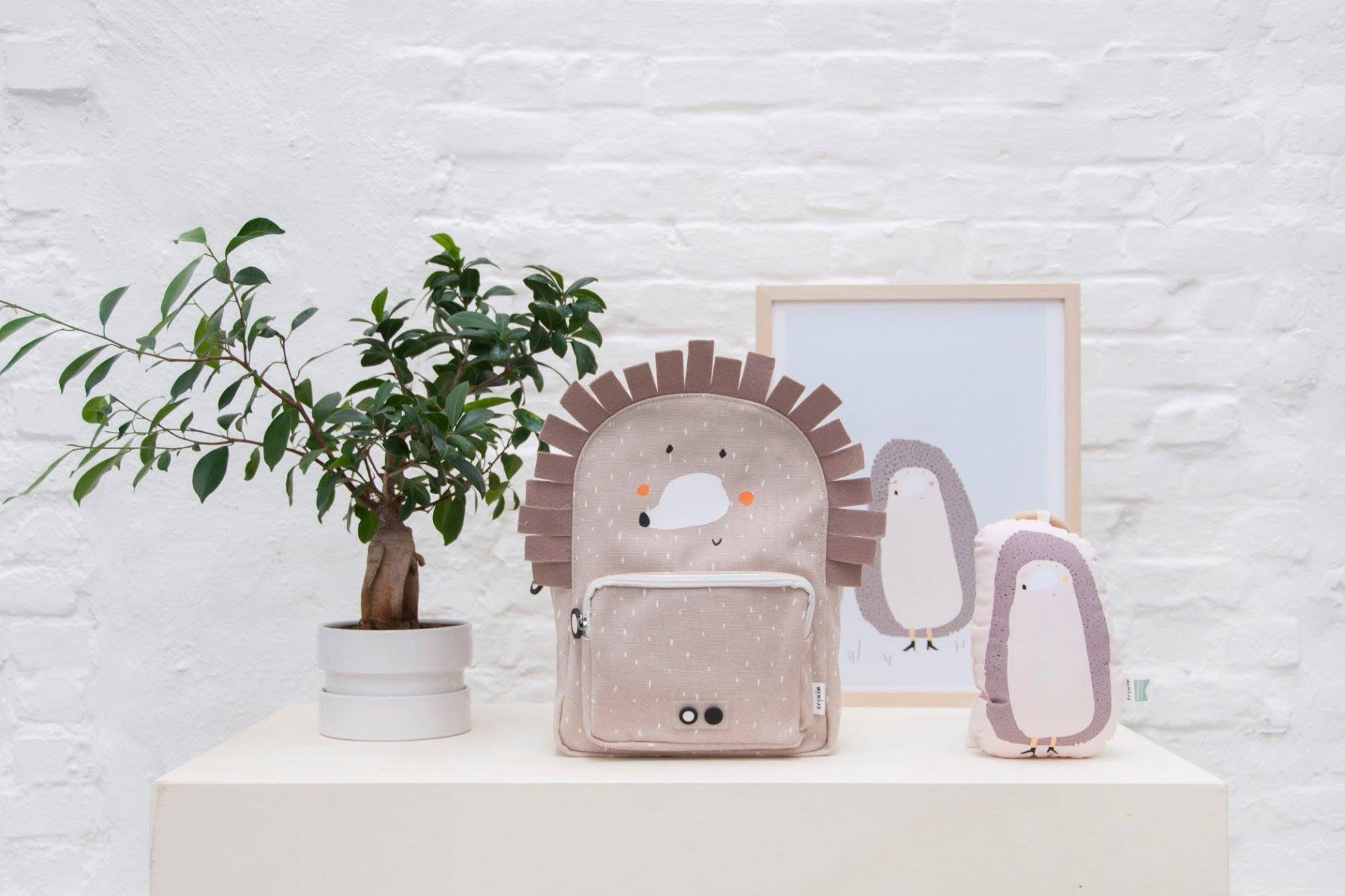 mochila infantil rosa claro com design de porco-espinho, bolso frontal, chaleira e quadro com ilustração de pinguim sobre mesa branca