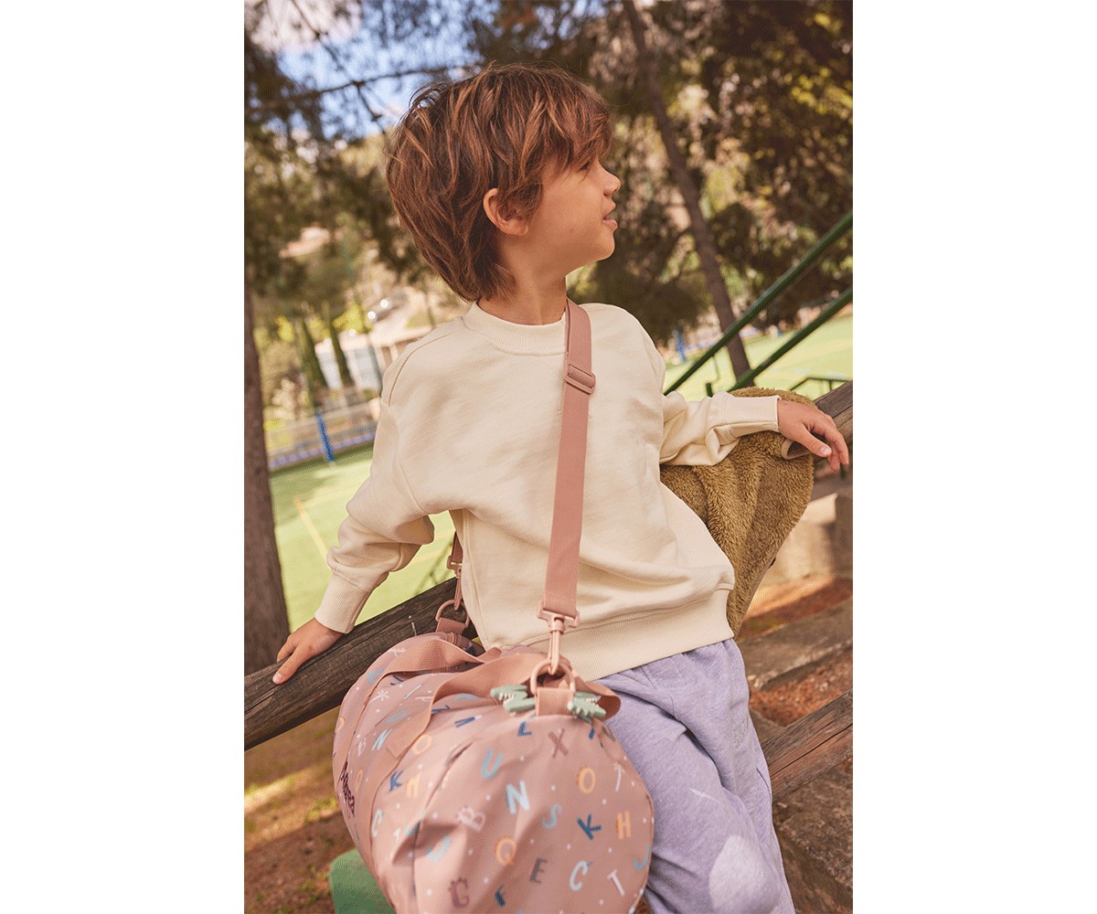 Criança com camisola bege e bolsa rosa com letras coloridas sentada num banco ao ar livre