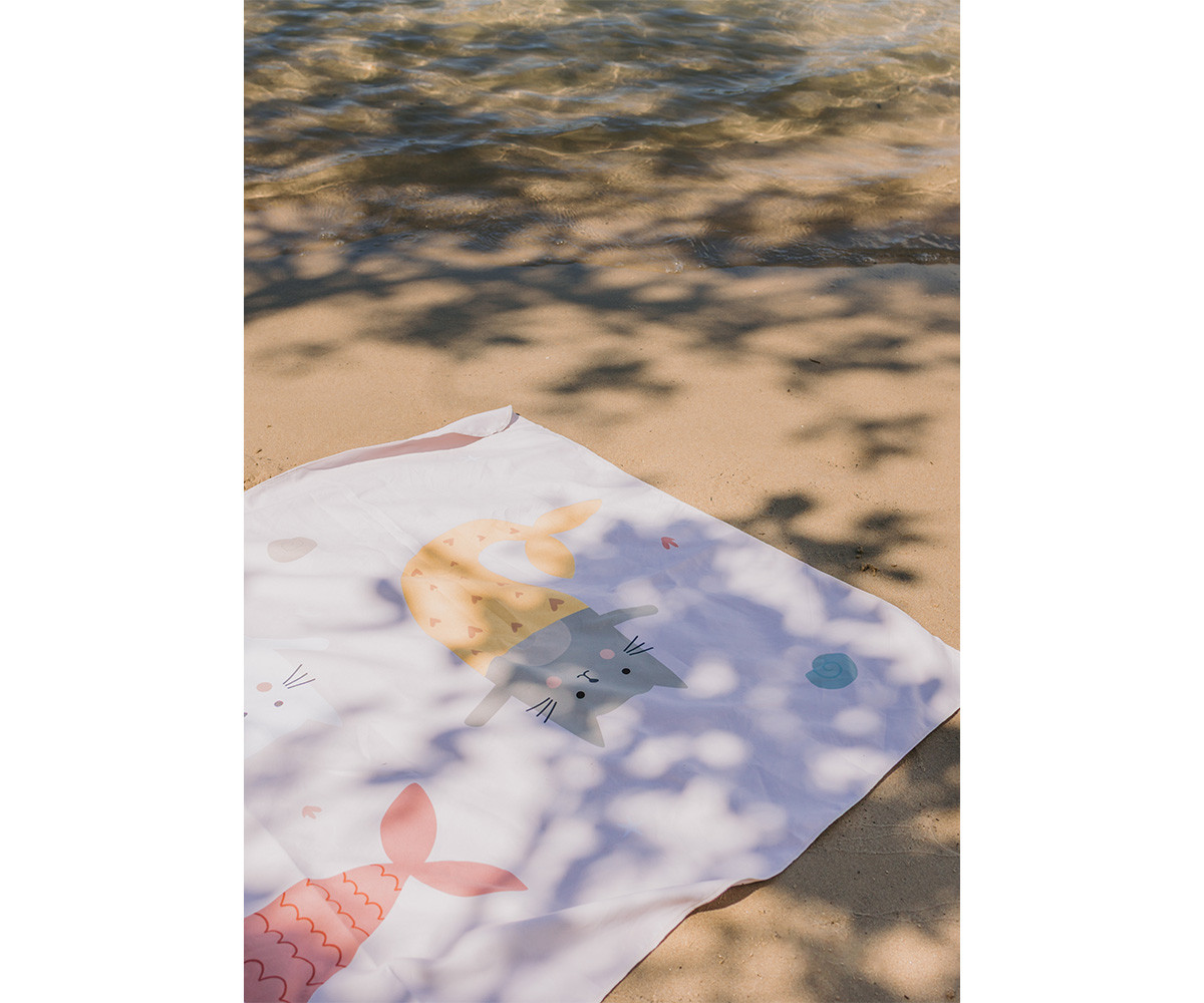 Paninho branco com desenhos de gatinhos sereias sobre areia e sombra de folhas