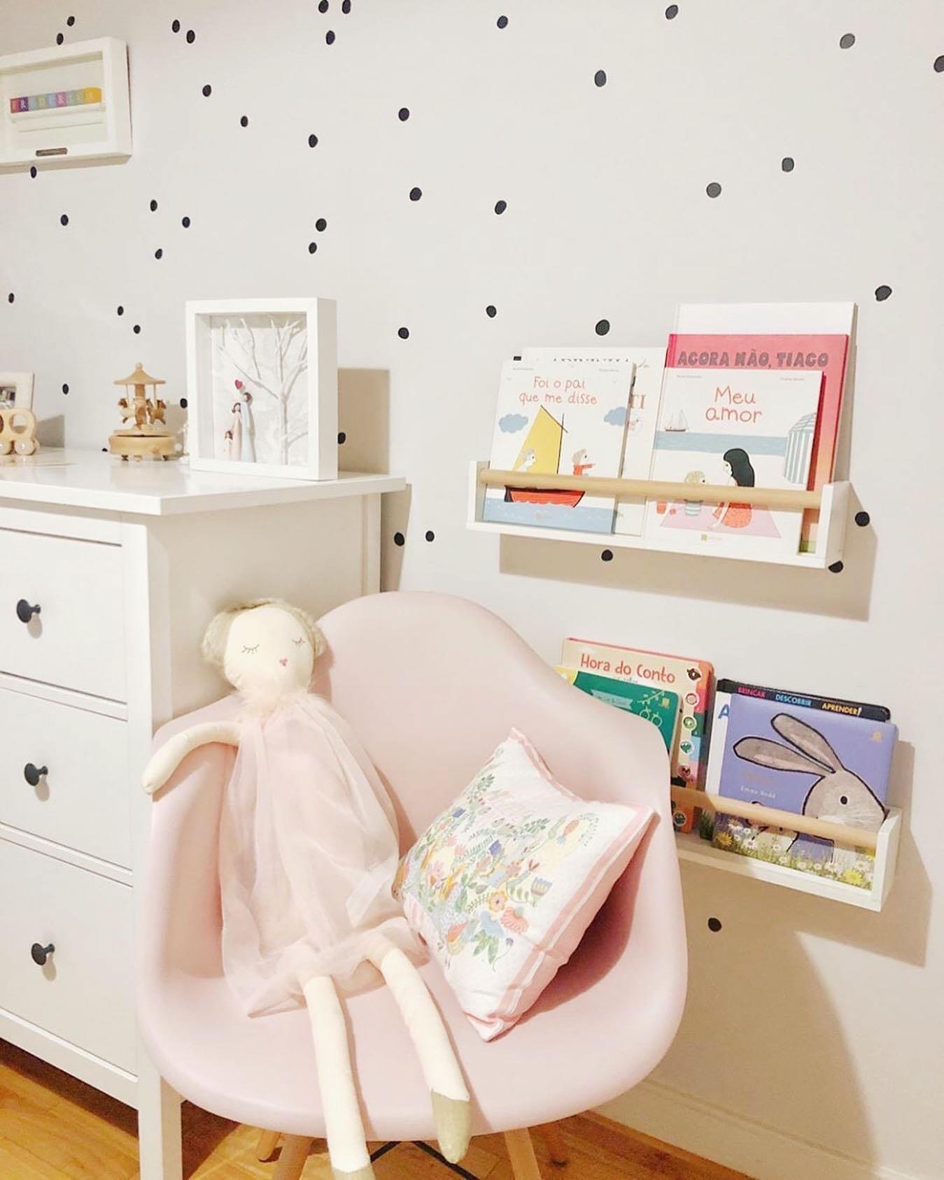 Cadeira rosa com boneca de pano, almofada e cómoda branca junto a parede decorada com pontos pretos e estantes com livros infantis.