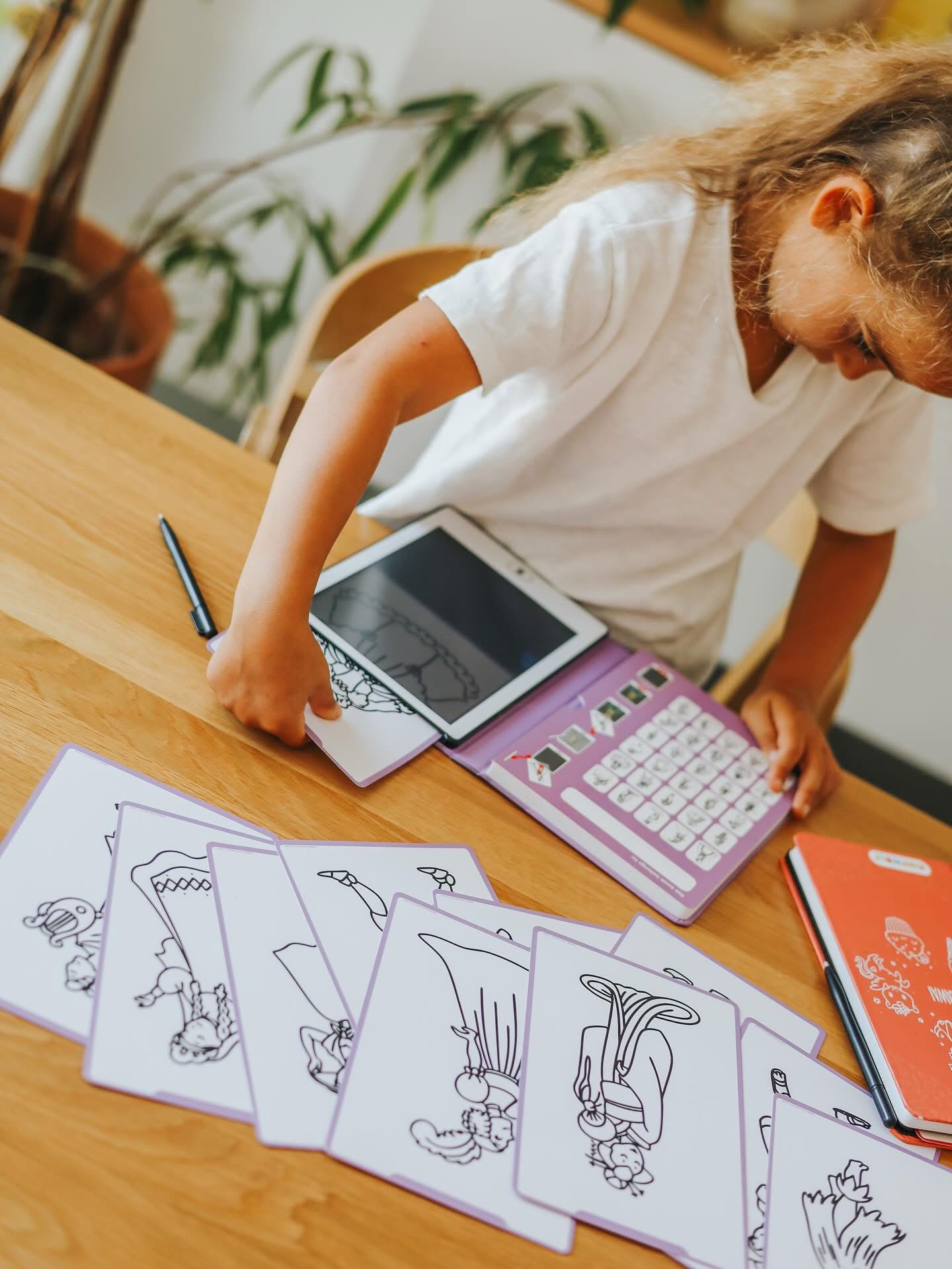 Criança usa tablet com teclado lilás e cartas de desenhos a preto e branco numa mesa de madeira.