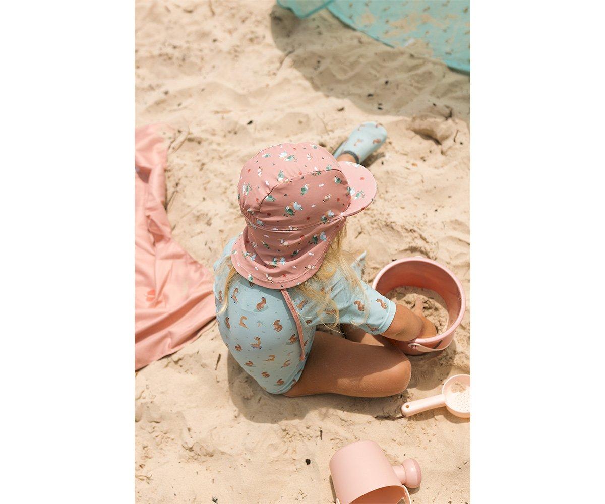 Criança com fato de banho azul e chapéu cor-de-rosa brinca com baldes e pás na areia da praia