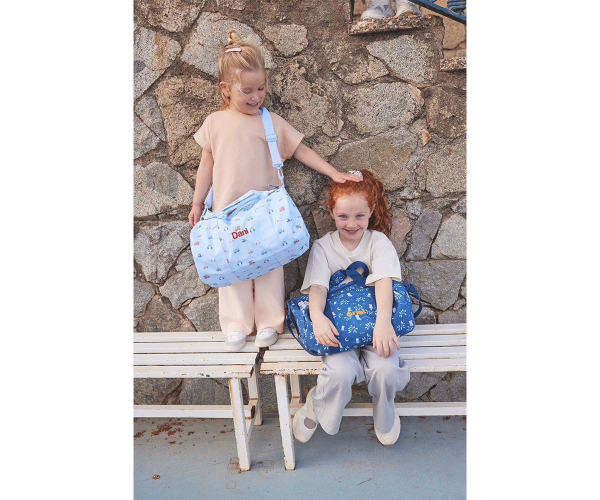 Duas crianças com sacos de maternidade azuis estampados sentadas e em pé em bancos brancos, parede de pedra ao fundo