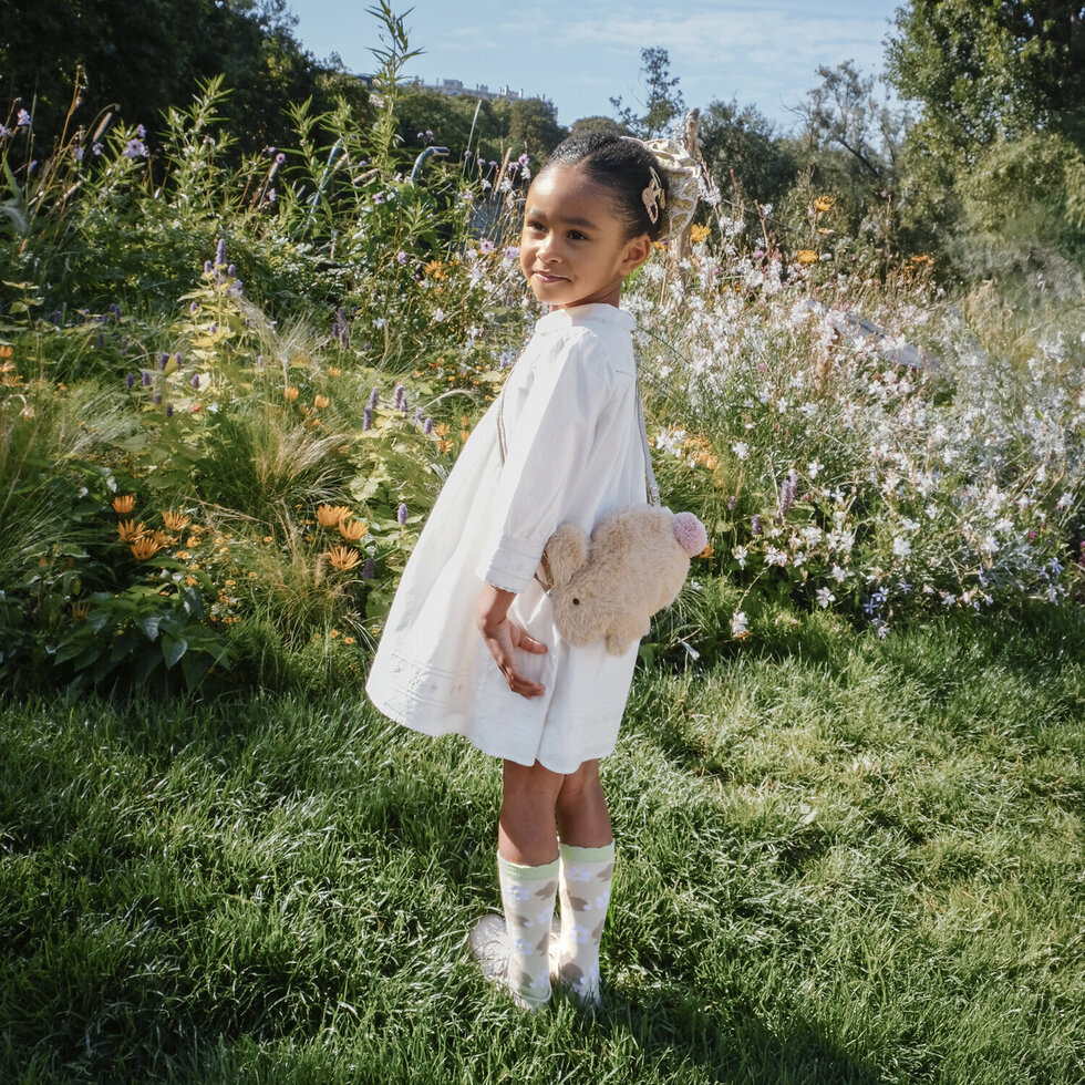 Menina com vestido branco, mala de peluche e botas transparentes em campo florido