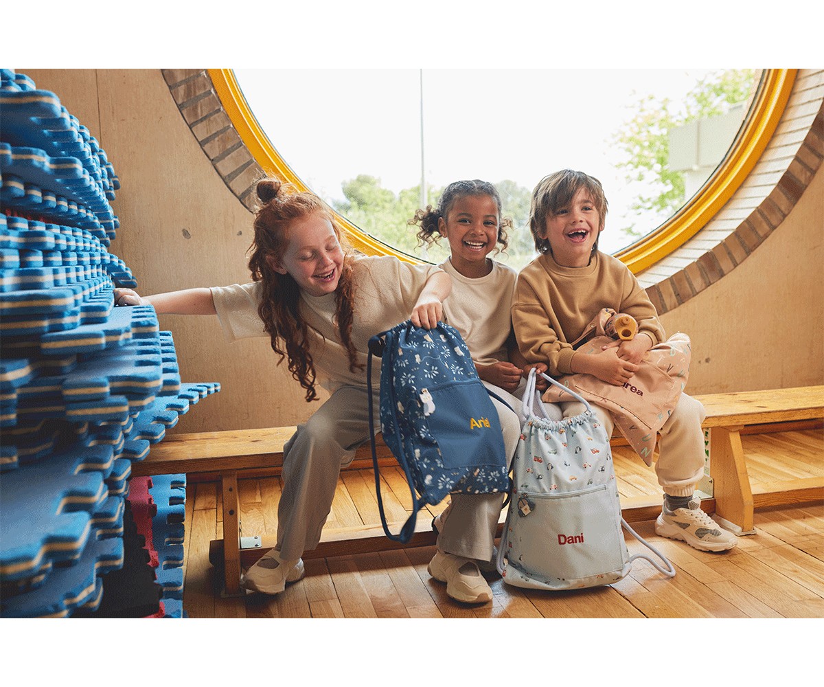 Três crianças sorridentes sentadas num banco de madeira junto a uma janela redonda, segurando mochilas personalizadas.