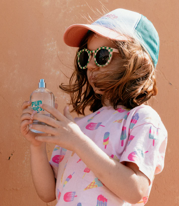 Menina com óculos quadrados, boné colorido e t-shirt de gelados segurando frasco spray transparente com texto azul