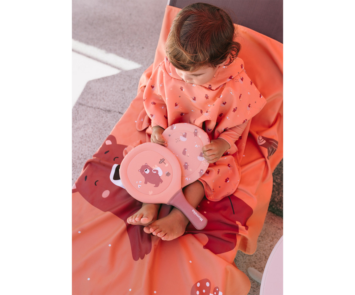 Criança com poncho e raquetes de praia laranja com desenhos de ursinho e cogumelos