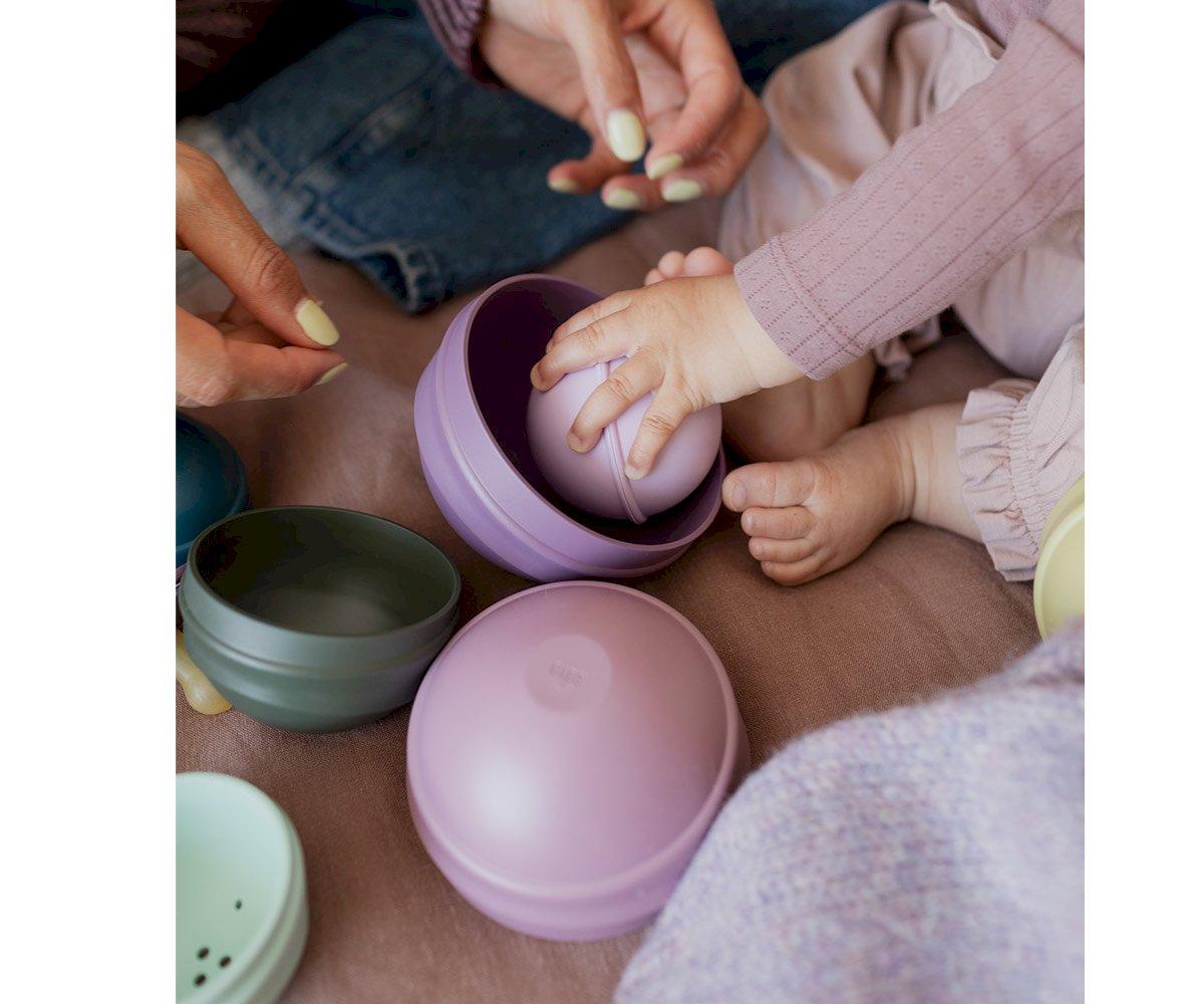 Mãos de adulto e criança a brincar com bolas e tigelas coloridas sobre superfície rosa