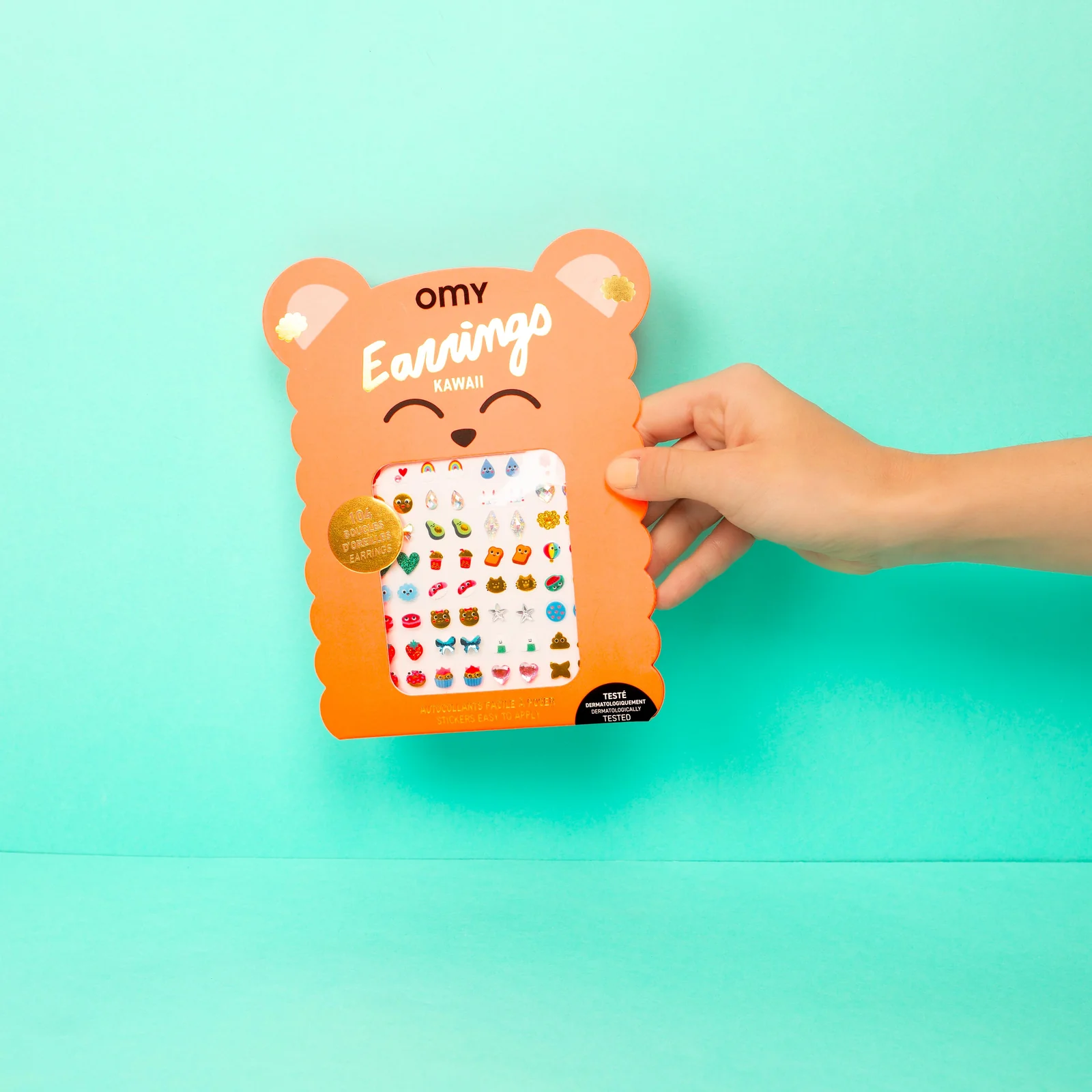 Embalagem laranja em forma de urso com brincos adesivos coloridos em fundo azul claro