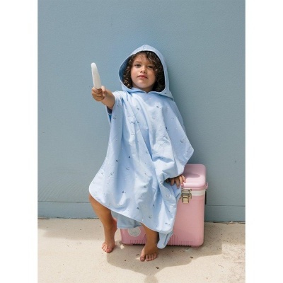 Criança com poncho azul claro e capuz, sentada numa mala térmica rosa, segurando gelado branco