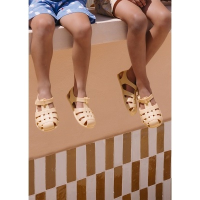 Dois pares de sandálias amarelas claras com design trançado, usadas por crianças sentadas numa parede com azulejos castanhos e brancos