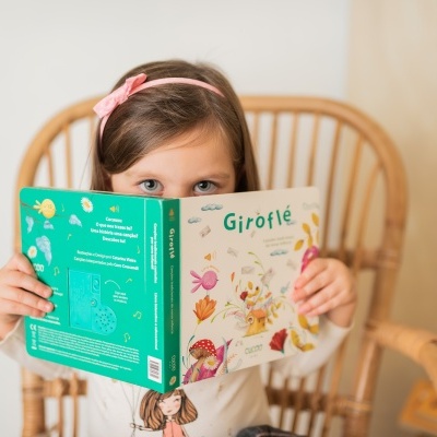 Criança a segurar um livro infantil 'Giroflé' com capa verde e branca, sentada numa cadeira de vime.