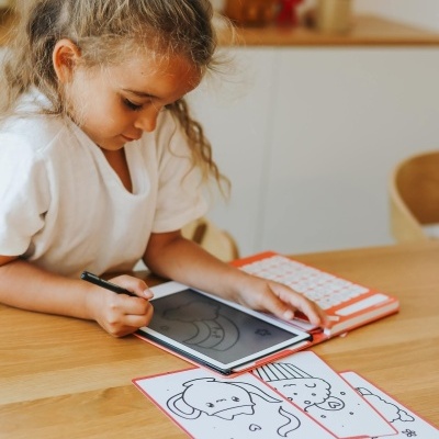 Criança a desenhar num tablet com capa laranja e teclado branco, mesa de madeira clara com desenhos infantis em preto e branco