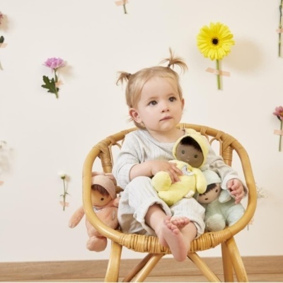 Bebé sentada numa cadeira de vime com três bonecos de tecido coloridos num quarto decorado com flores na parede